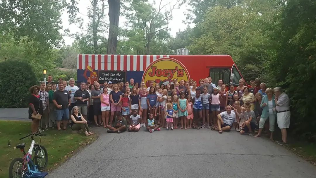 Large group of people posing in front of a red food truck with trees in the background.