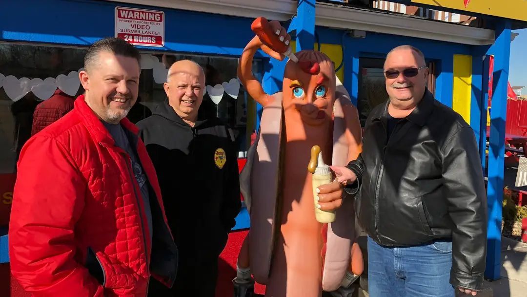 Four men posing with a hot dog mascot outside a food stand; sunny day.