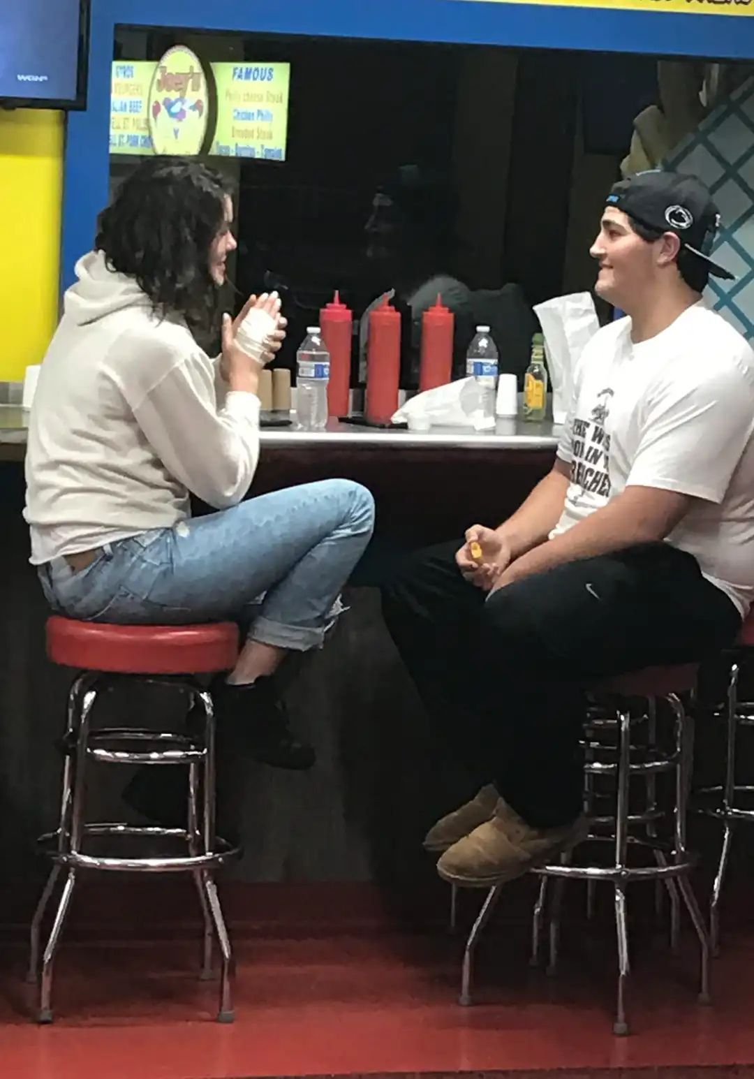 Two people talking, sitting at a counter with condiments and water bottles. One wears a hoodie, jeans. The other wears a t-shirt, cap, and sweatpants.