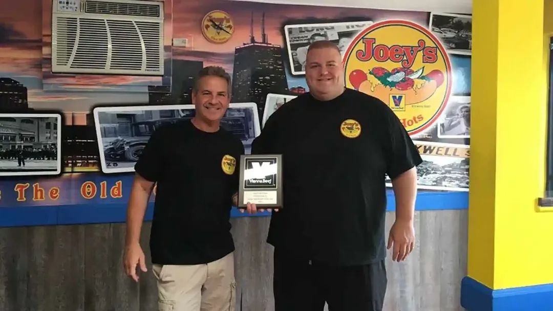 Two men holding an award in front of a Joey's Hot Dog sign in a diner.