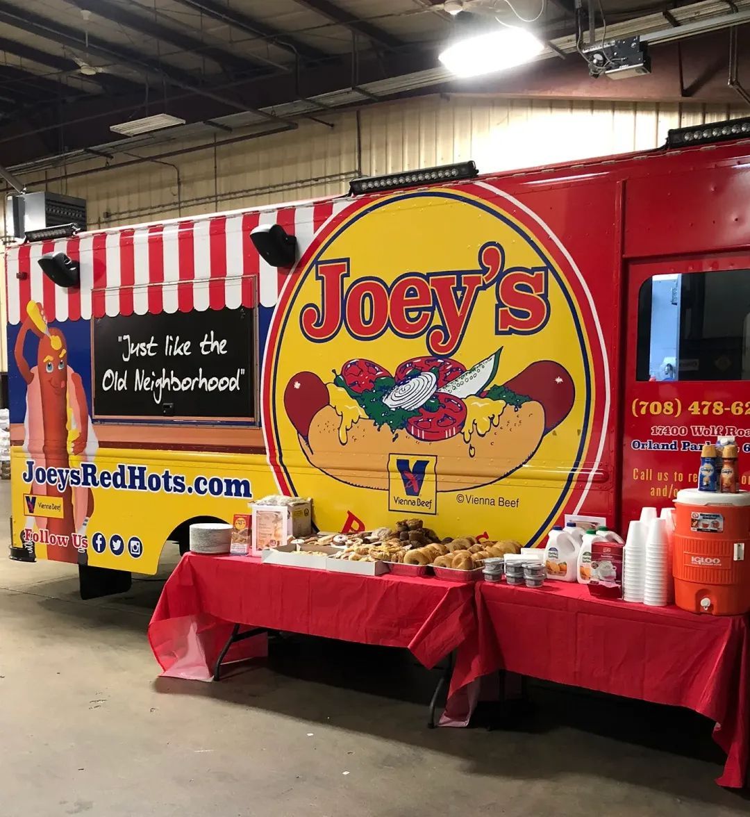 Joey's Red Hots food truck with a red and yellow logo. Red table with food and drinks set up in front of the truck.