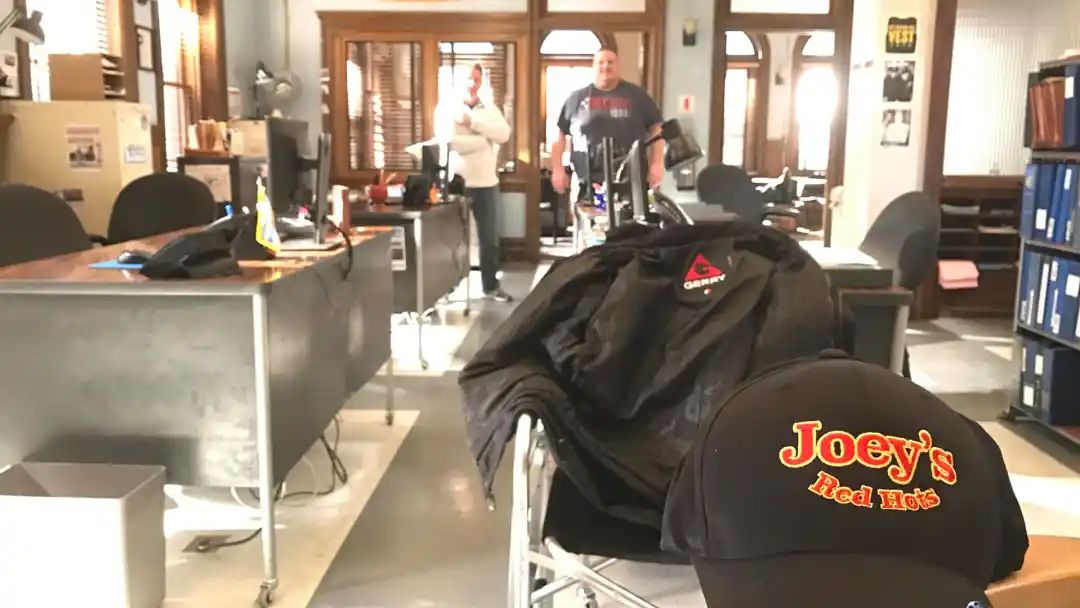 Office with two people, desk, black jacket and Joey's Deli hat on a chair.