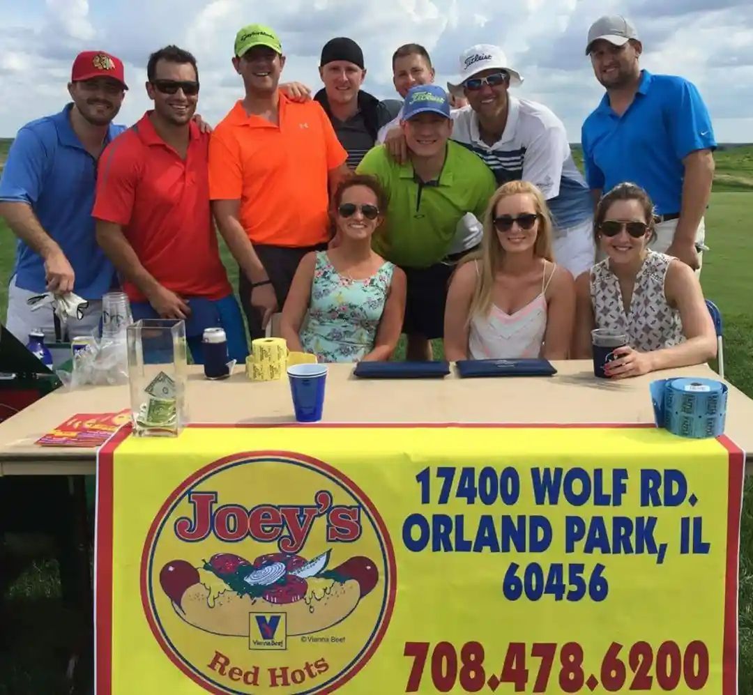 Group of people with Joey's Red Hots stand on a golf course. Some are smiling, and the stand displays the address and phone number.