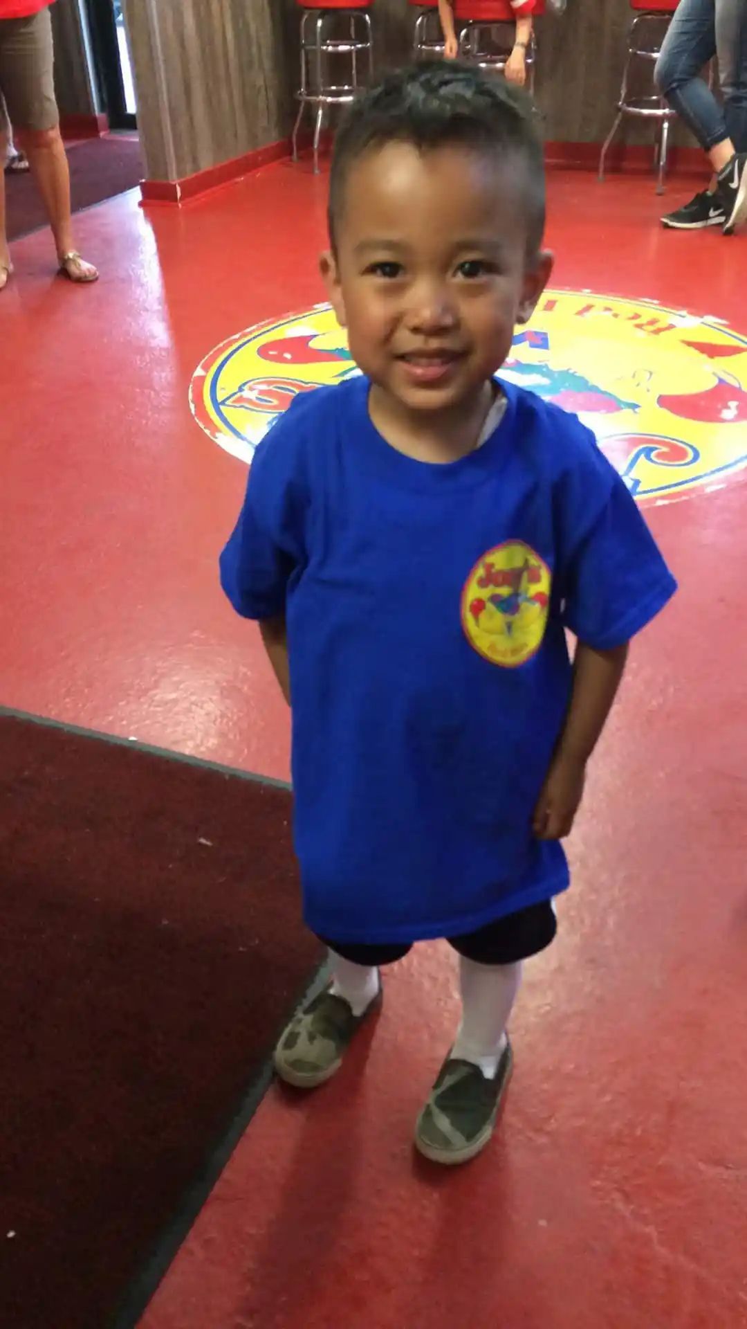 Boy in a blue shirt with logo, black shorts, and green shoes stands on red floor, smiling.