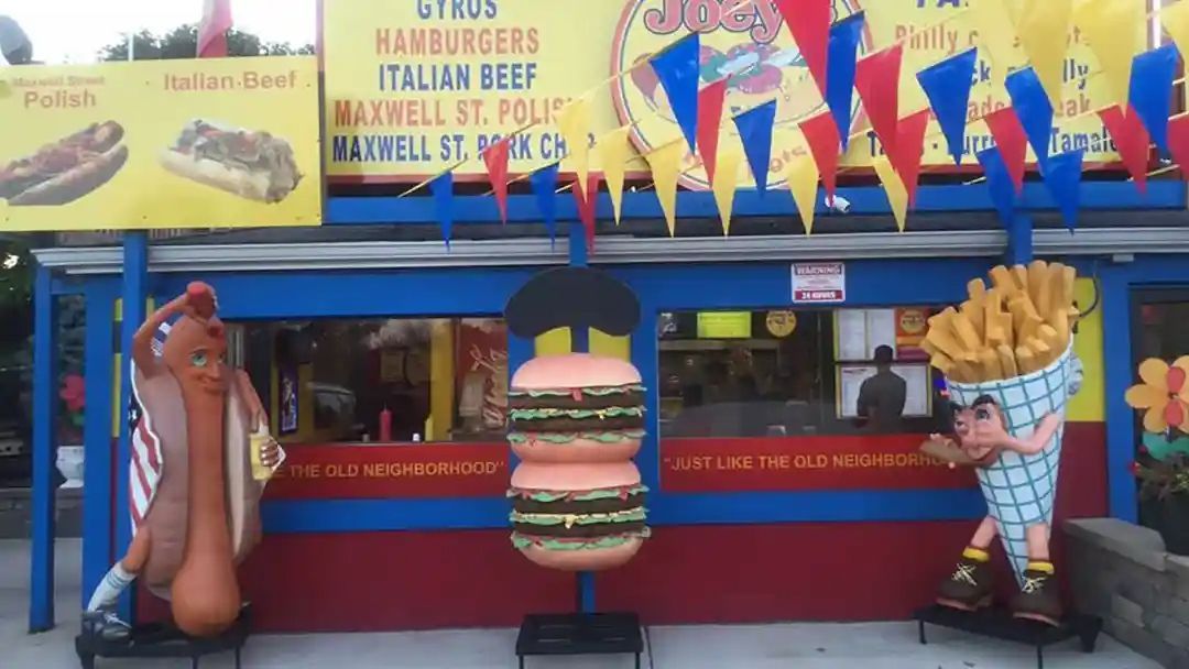 Food stand with giant hot dog, burger, and fries sculptures. Blue and red facade with food menu signs.