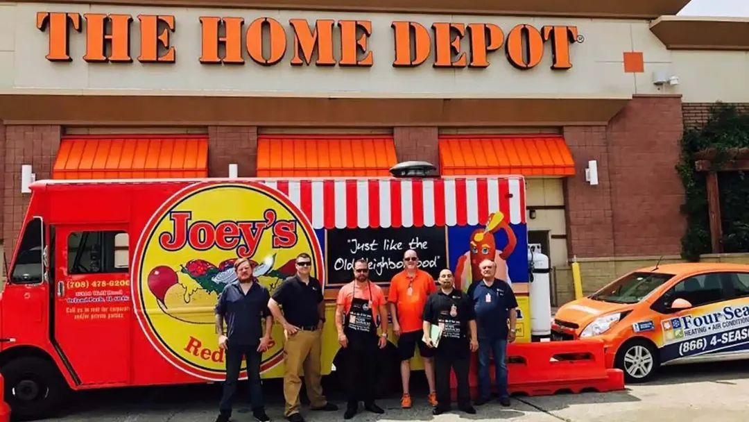 People stand in front of a Home Depot and food truck, Joey's Red Hots. Orange and red colors are prominent.