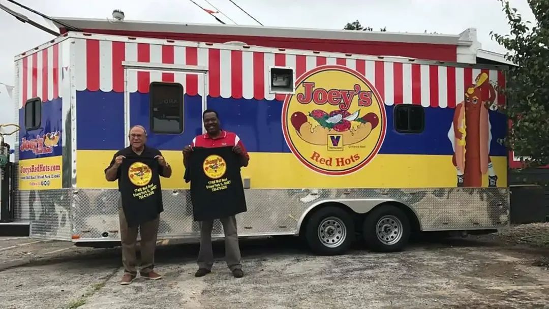 Two men holding t-shirts in front of a colorful food trailer, “Tony’s Hot Dogs.”