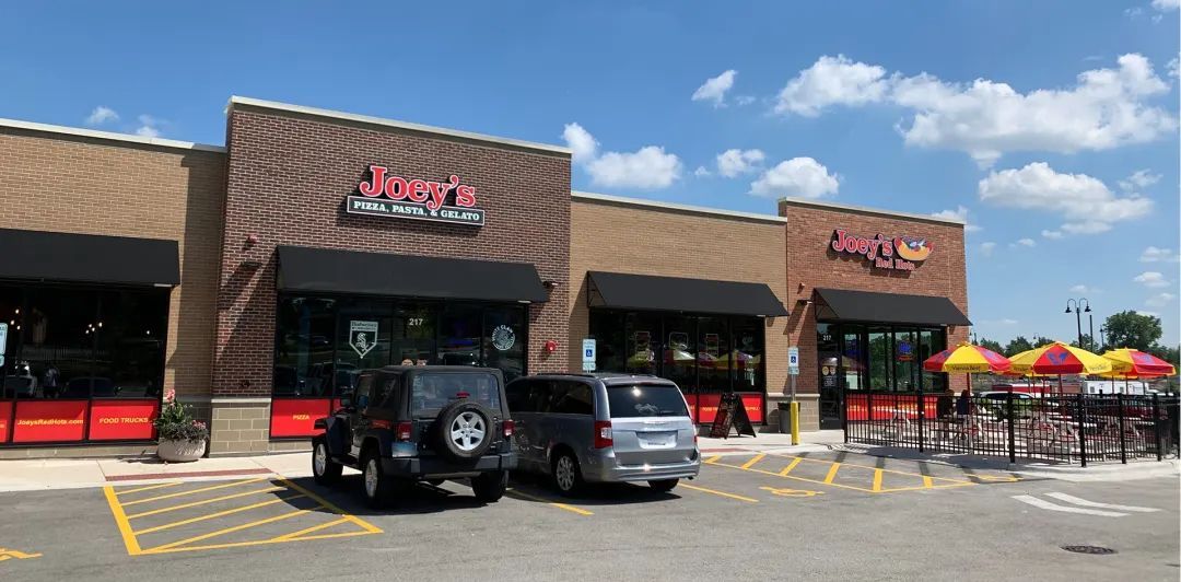A strip mall with two restaurants, Joey's, and an unidentified one. Cars parked in front with outdoor seating.