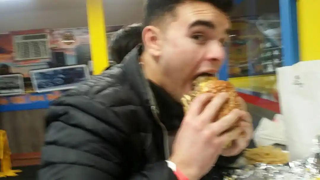 Man taking a large bite of a burger indoors, wearing a black jacket; yellow and blue wall in the background.
