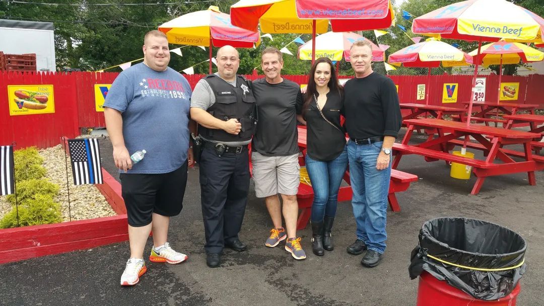Five people pose outside a red hot dog stand with umbrellas and picnic tables.