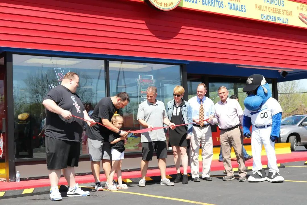 People cutting a ribbon at a restaurant opening, including a mascot. Red building, sunny day.