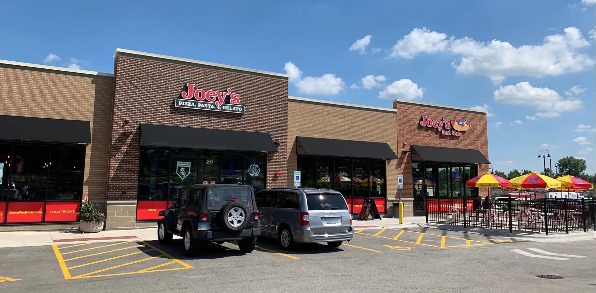 Exterior of two Jimmy John's stores with cars parked in front and outdoor seating with umbrellas.