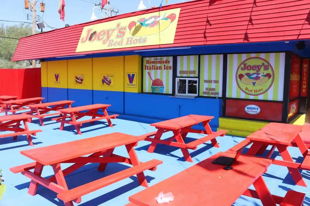 Joey's Red Hots food stand with red picnic tables on a blue patio. Yellow and blue building with red roof.