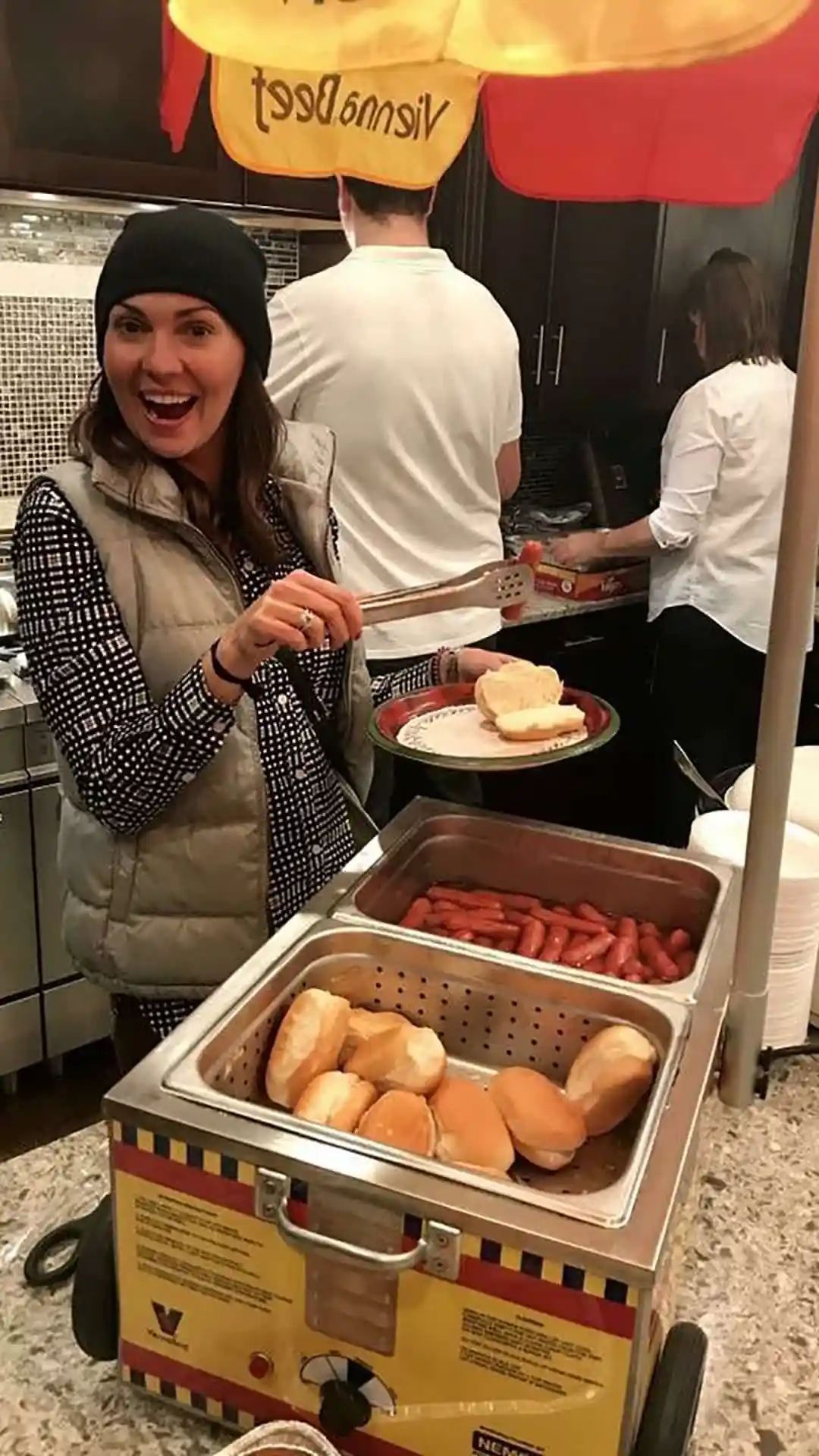 Woman with tongs near hot dog cart, smiling in kitchen. Hot dogs, buns, and yellow umbrella visible.
