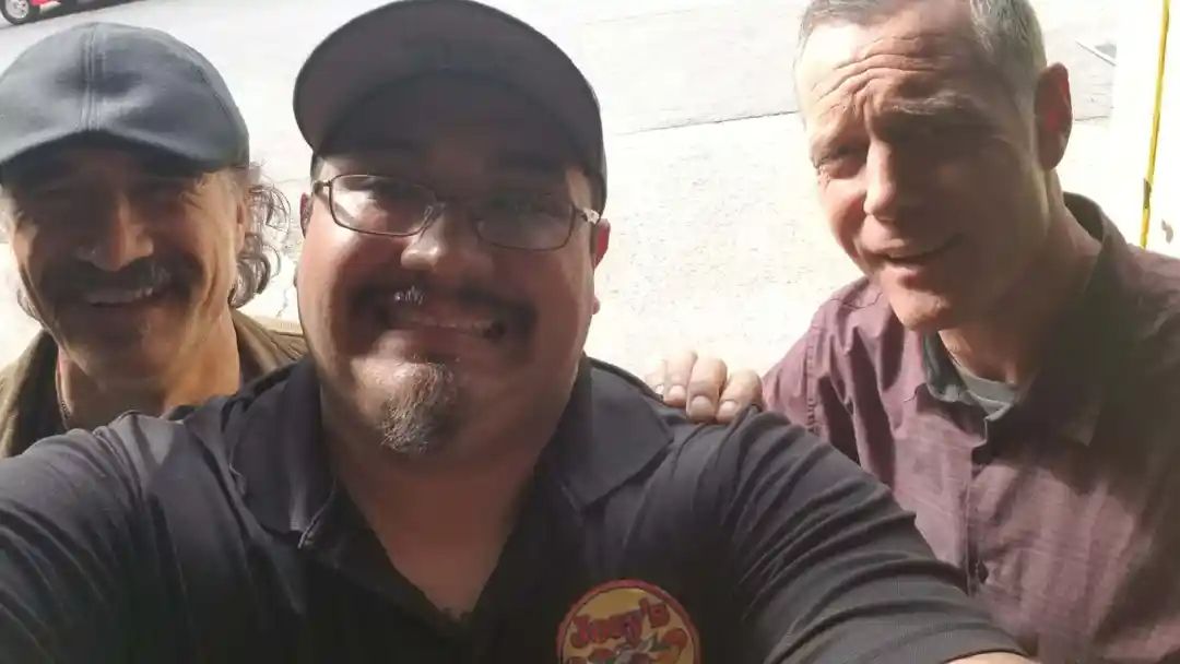 Three men smiling for a selfie; the central man has a beard and glasses, wearing a cap.