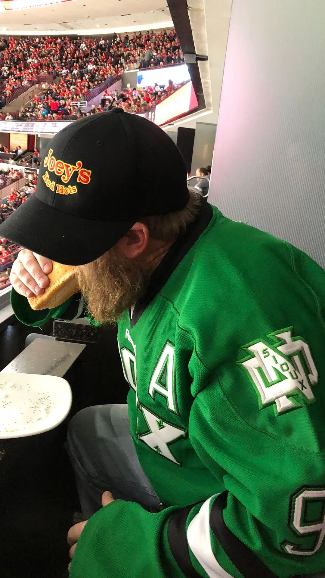Man in Dallas Stars jersey eating at a hockey game, wearing a hat with text, stadium in background.