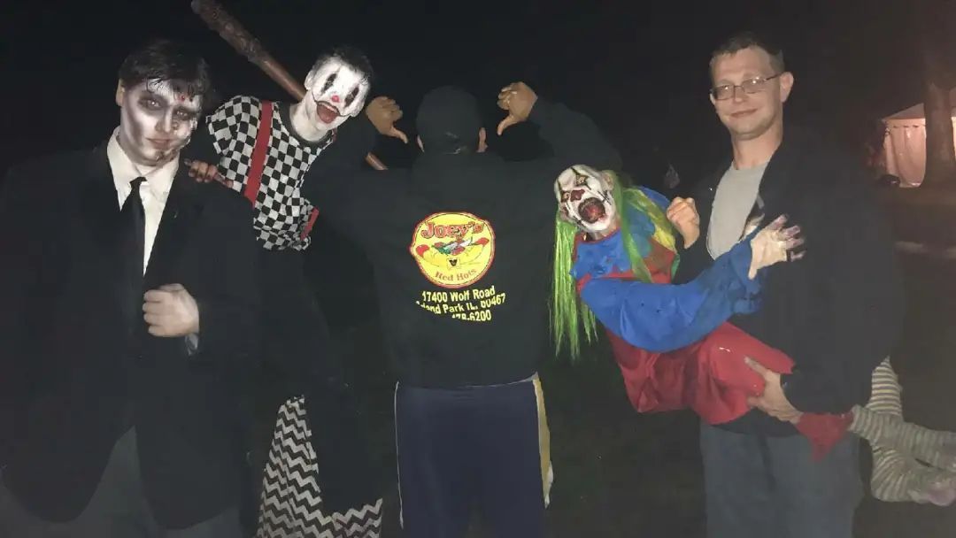 Five people in costumes pose at night. Two are dressed as clowns, one holding another clown. Others wear scary makeup.