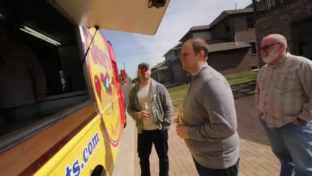 Three men standing at the window of a yellow food truck on a sunny day.