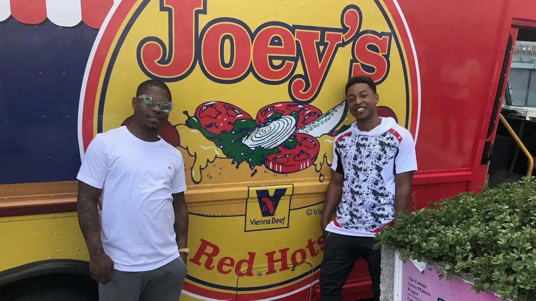 Two men stand in front of Joey's Red Hot food truck with yellow, red, and green imagery.