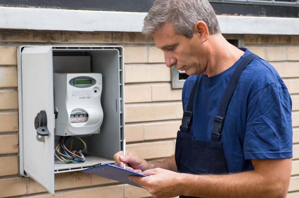 A Man is Looking at a Meter in a Box While Holding a Clipboard — AES Electrical Contracting In Winnellie, NT