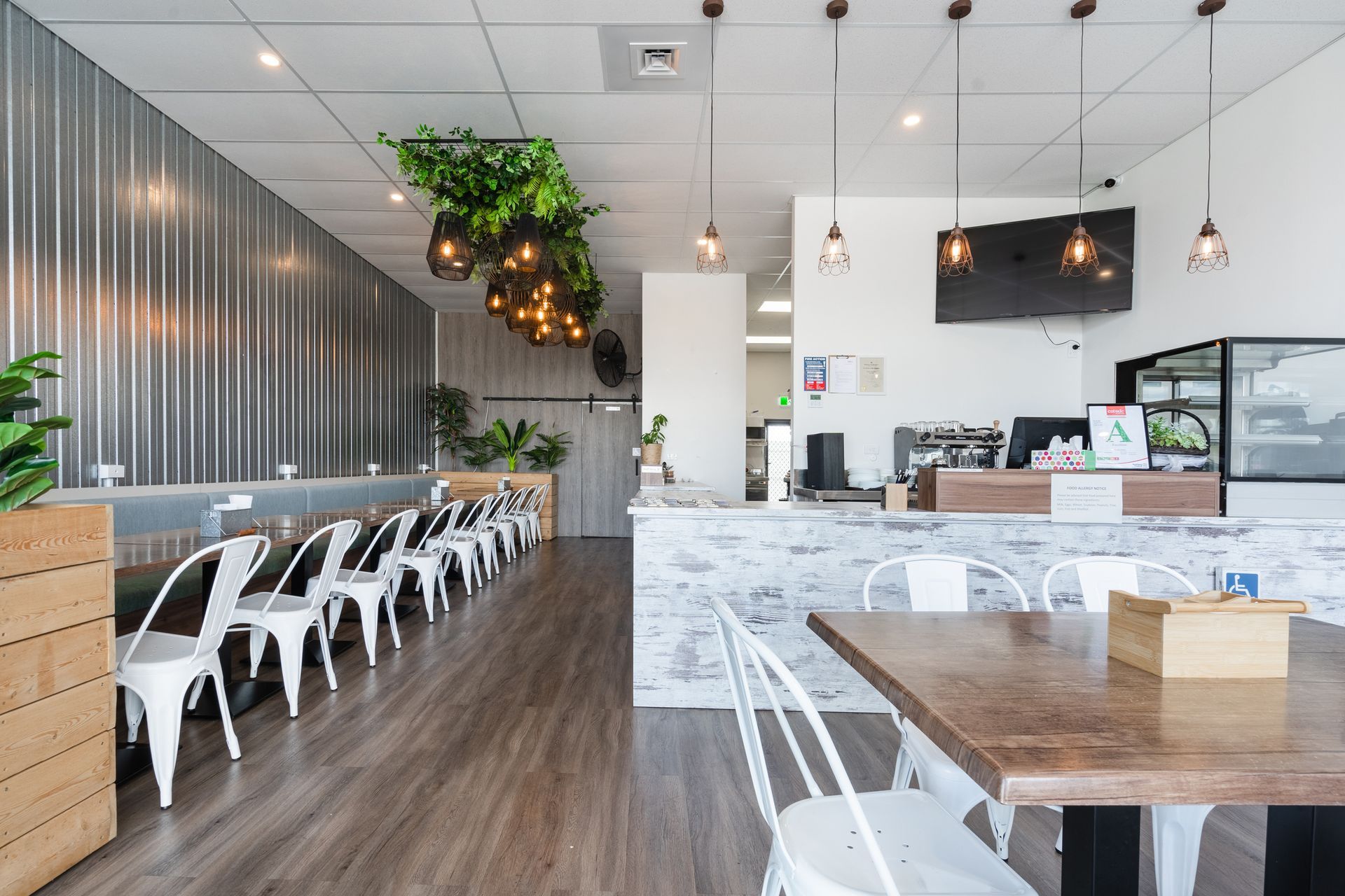 Modern café interior with wooden floors, white chairs, hanging lights, and green plants.