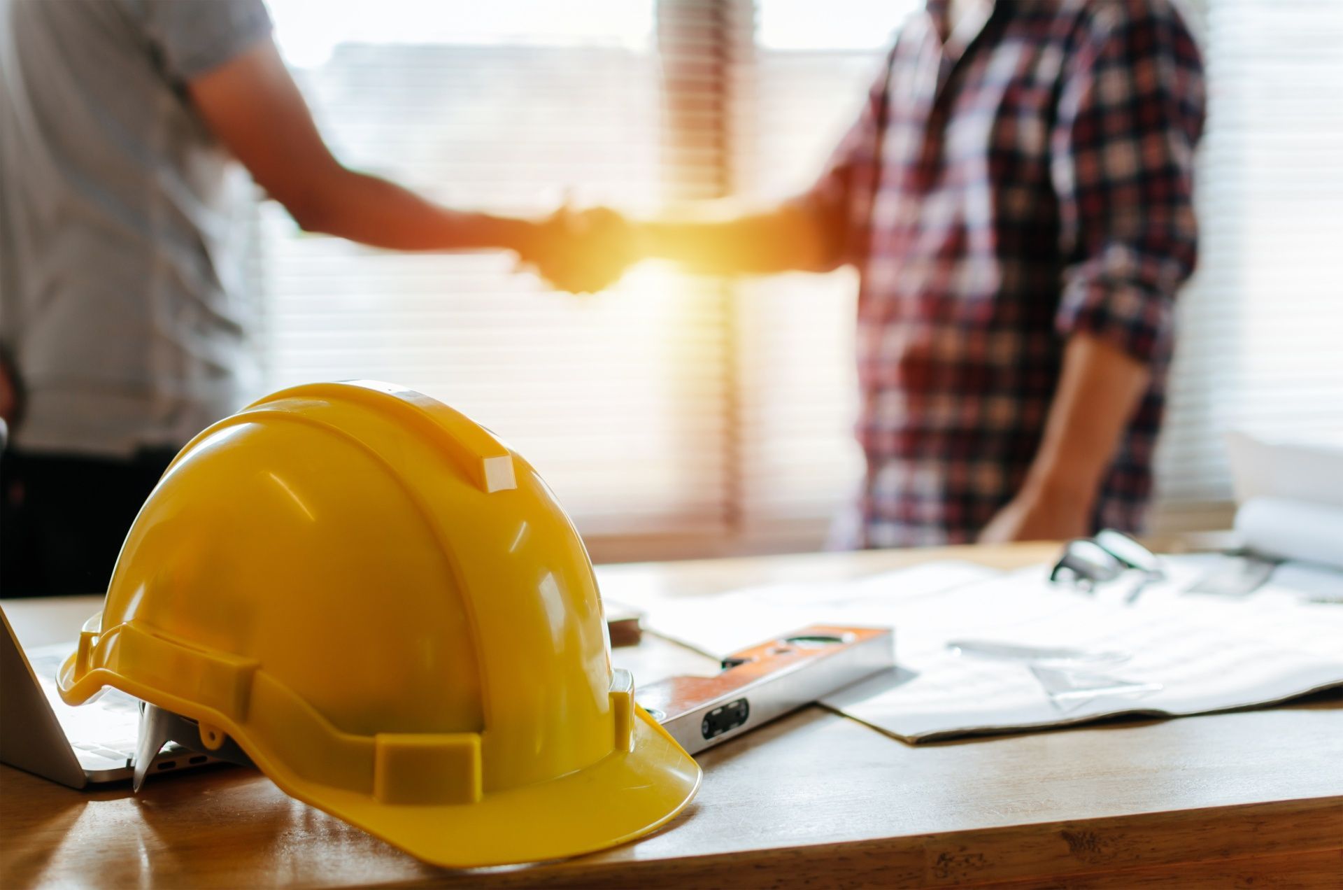 Two men are shaking hands over a table with a hard hat on it.