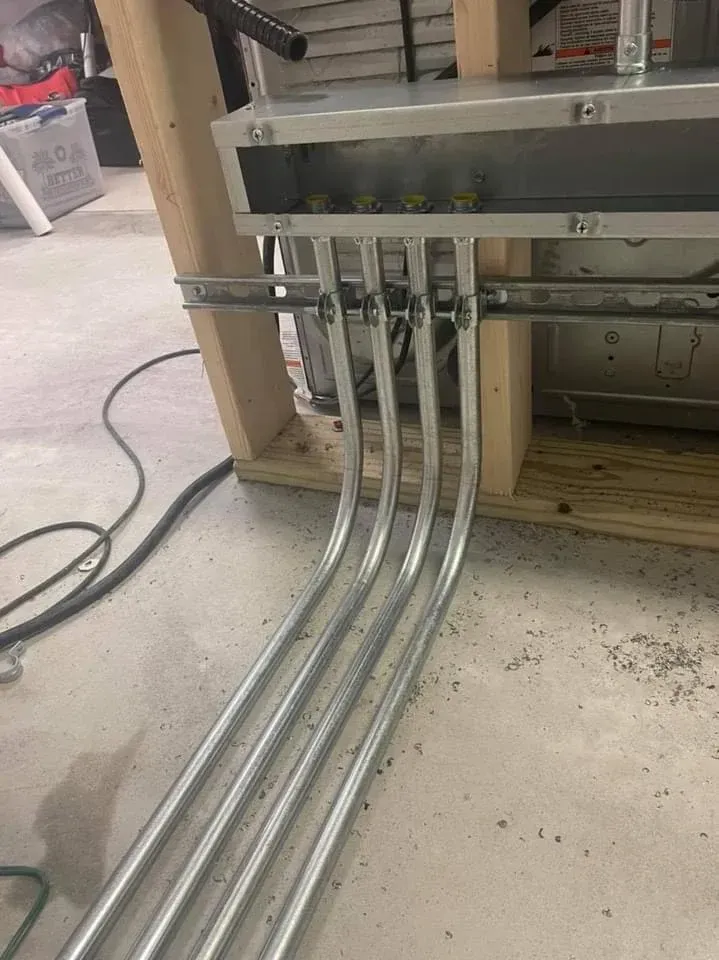 Four metal conduits curving upward into an electrical box, mounted on wood, set on a concrete floor.