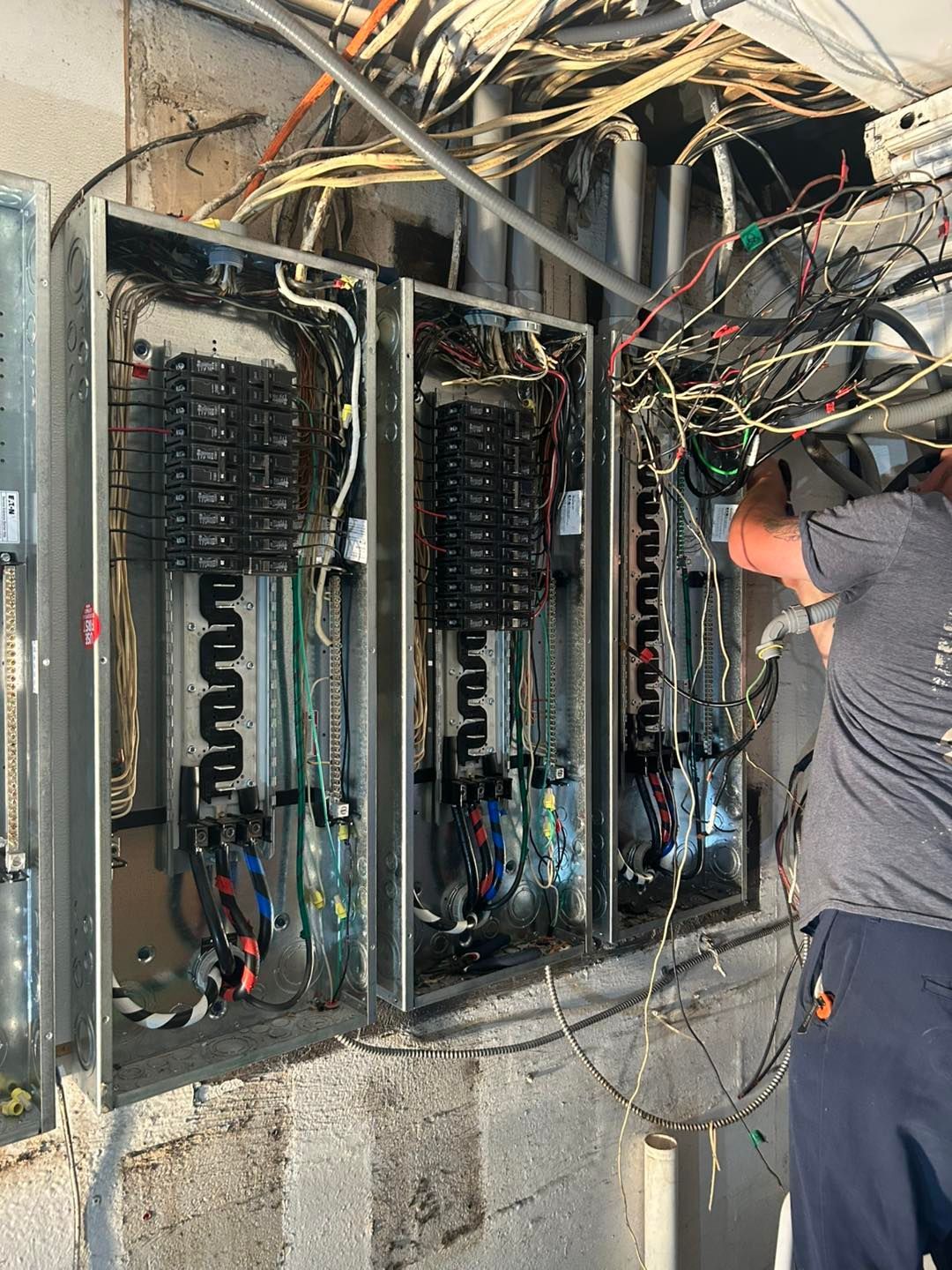 Electrician working on exposed electrical panels with tangled wires in a building setting.