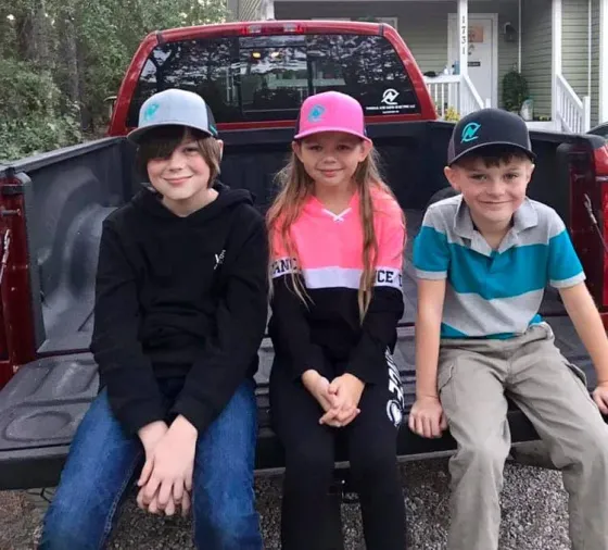 Three children in hats sit on a truck bed in front of a house.