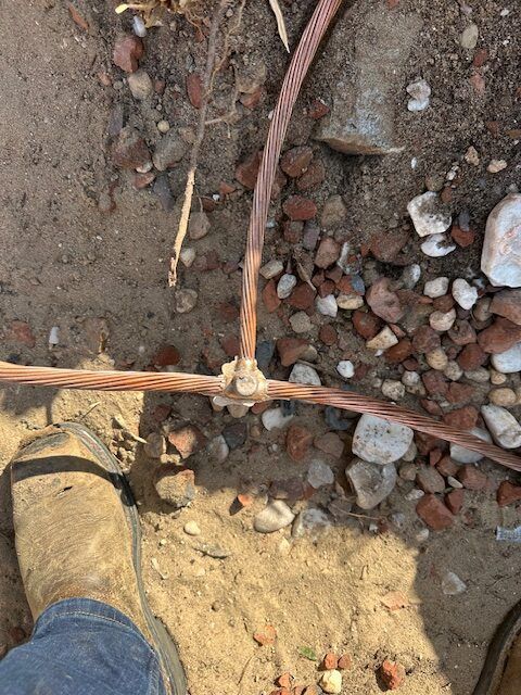 Copper wires connected with a clamp on dirt and rocks, viewed from above near a boot.