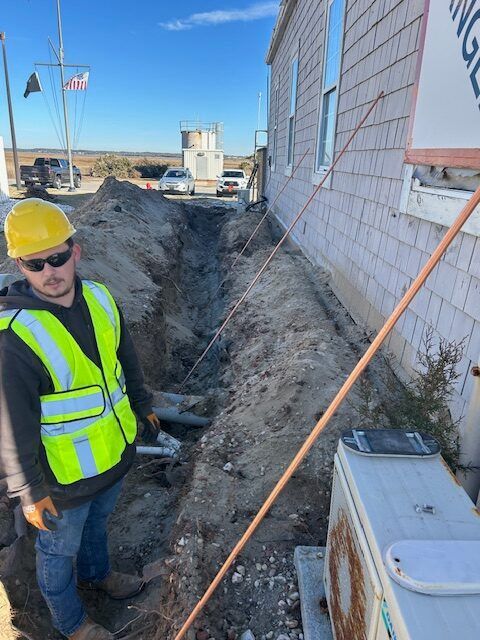 Construction worker in safety gear by a trench next to a building. Sunny outdoor setting.