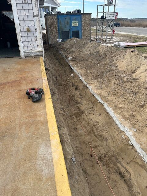 Trench dug beside a building, dirt pile, construction site. Ground is sandy and yellow border. Red equipment in foreground.