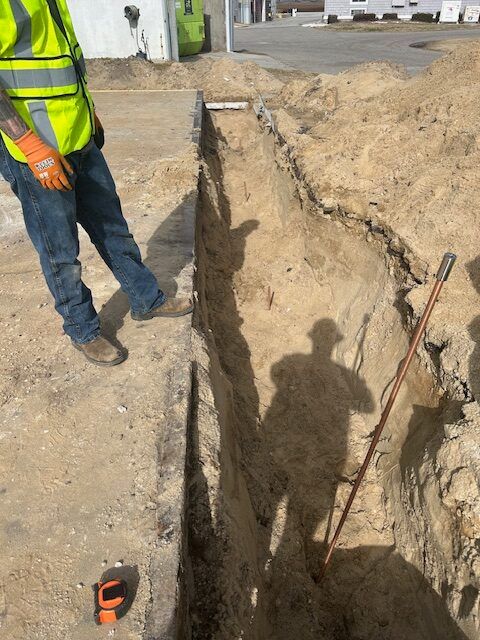 Person in safety vest near a trench in dirt.  Shadow visible. Sunny outdoor construction site.