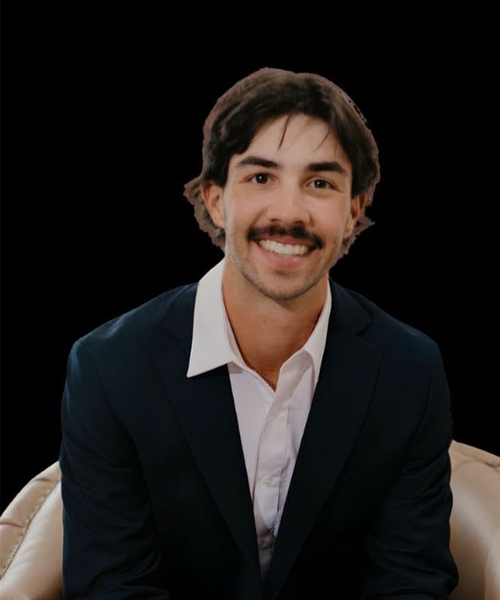 A smiling person with dark hair and a mustache wearing a dark suit and white collared shirt, seated in a beige chair.