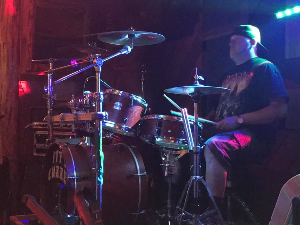 Drummer playing a drum kit under colorful stage lights. The person wears a cap and is in a dark room.