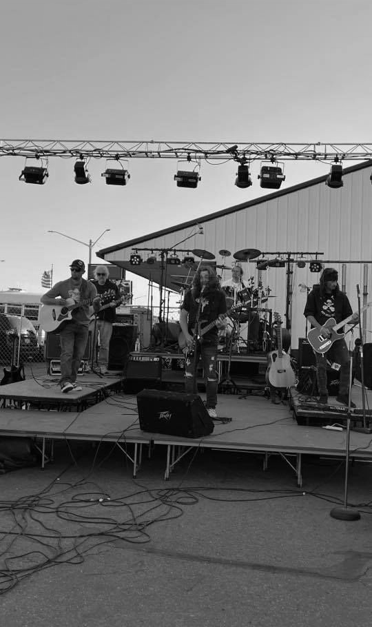 Band performing on an outdoor stage with guitars, drums, and stage lights.