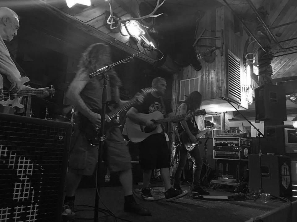 Band performing on stage, playing guitars, bass, and drums. Dimly lit bar setting.