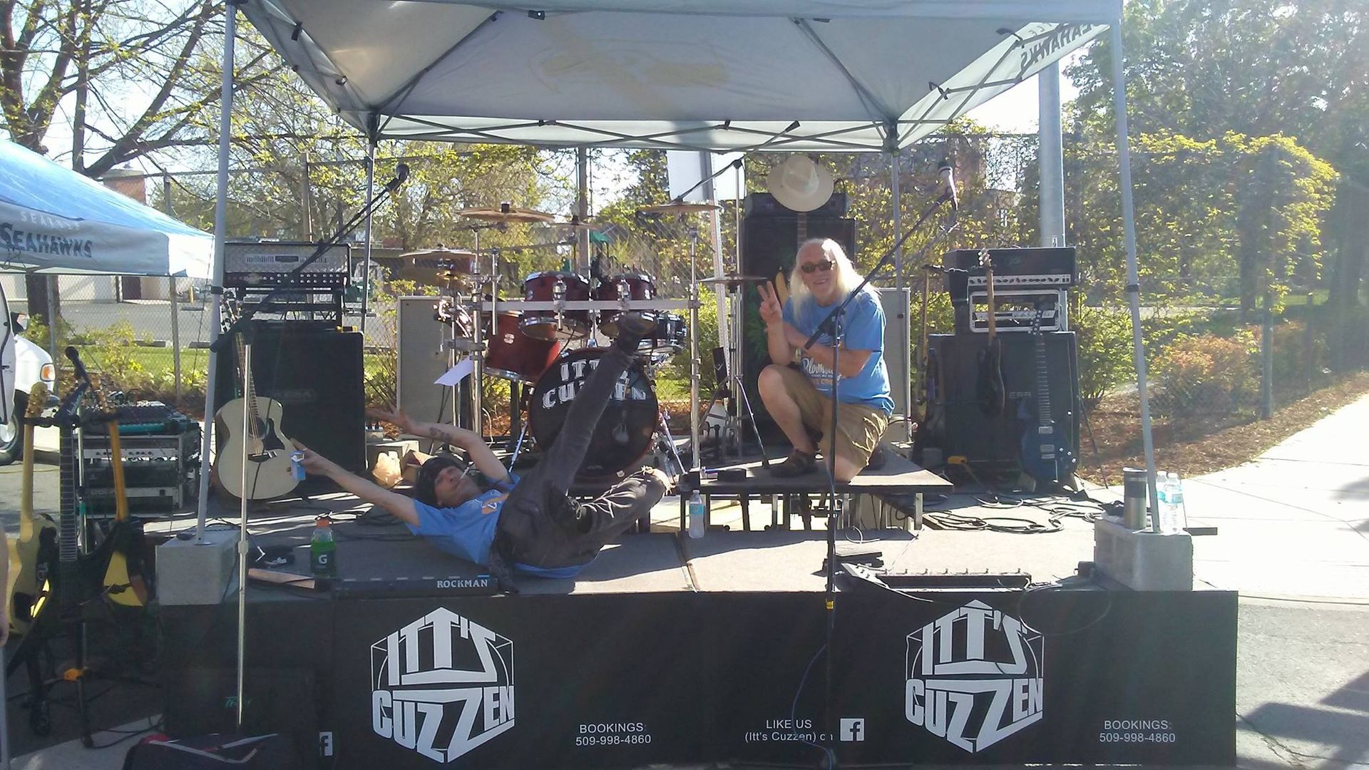 Band performing on outdoor stage; drummer and another person on the stage, one person on ground with arms up.