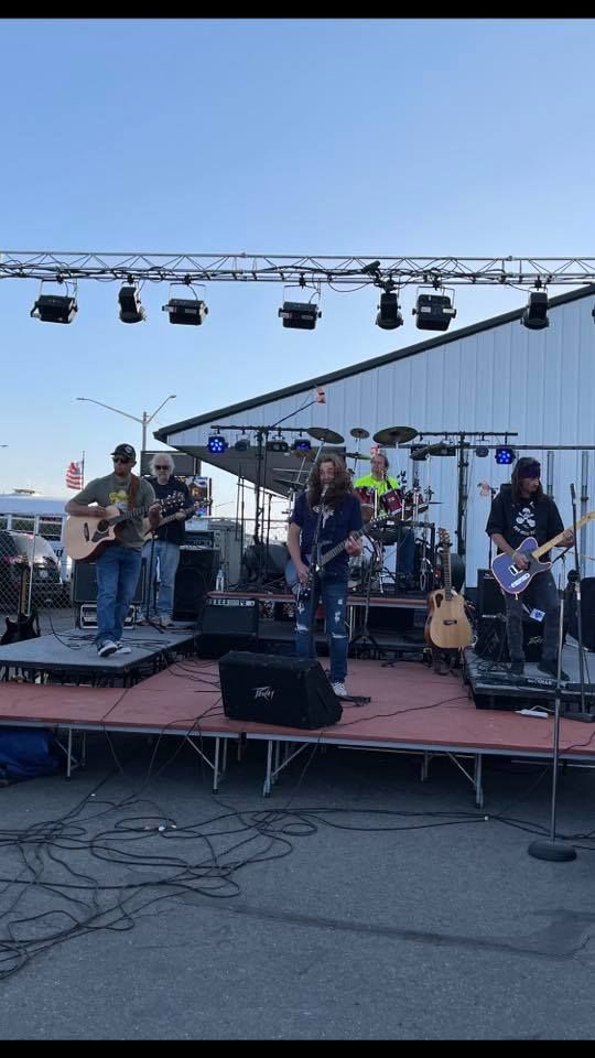 Band performing outdoors on a stage with a lighting rig. Musicians playing guitars, drums, and singing.