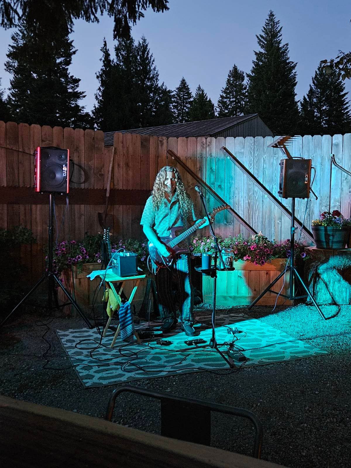 Person playing electric guitar on a small stage with blue and pink lights in a backyard.
