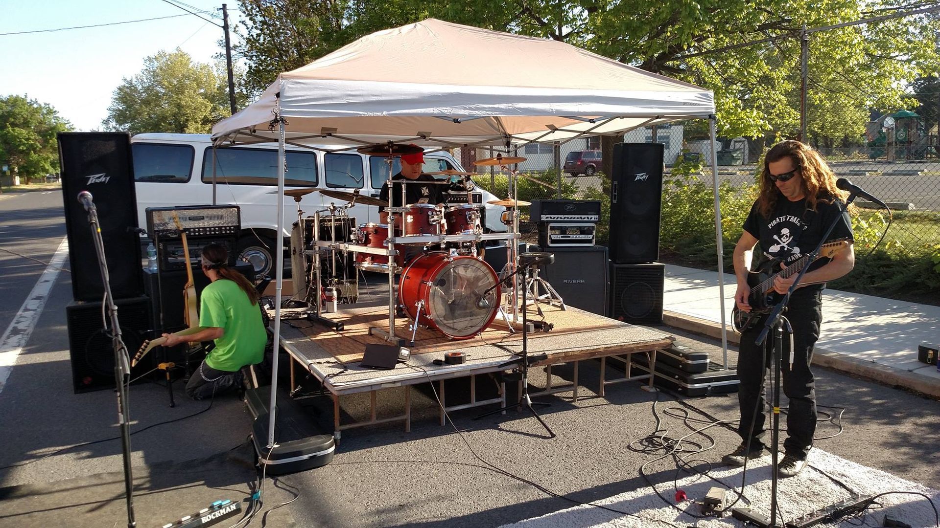 A band playing outdoors on a makeshift stage. Drummer in the center, guitarist right, another member on the left.