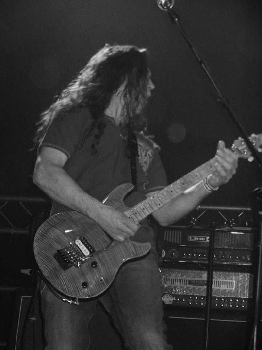 Guitarist with long hair playing an electric guitar onstage under a microphone.