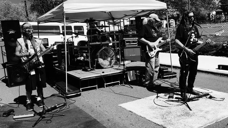 Band performing outdoors under a white canopy. Drummer, bassist, and guitarist play on a stage in grayscale.