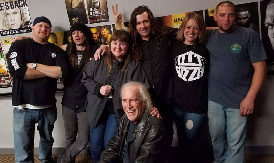Group of people smiling, posing with a smiling man seated in front. Movie posters are in the background.