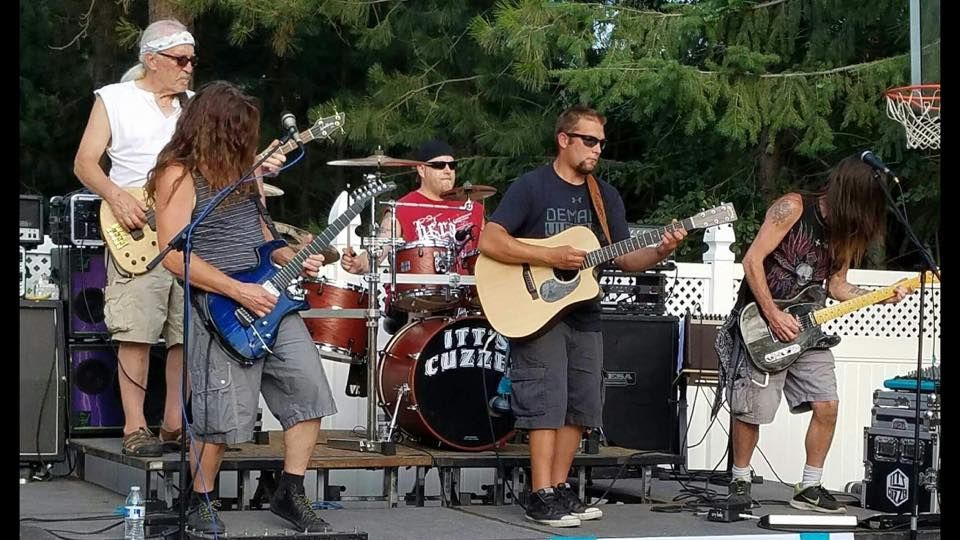 Band playing on a stage outdoors: drums, guitars, and a singer, performing in front of a green backdrop.
