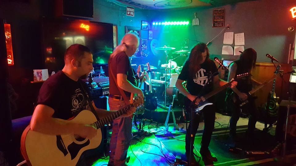 Band playing live in a bar; guitarist, bassist, drummer, and a singer under green and blue stage lights.