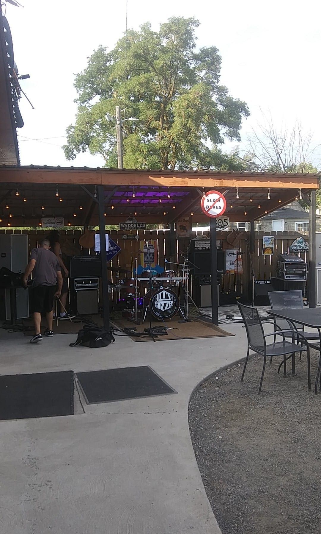 A band is setting up on a covered outdoor patio. There are drums, amps, and sound equipment.