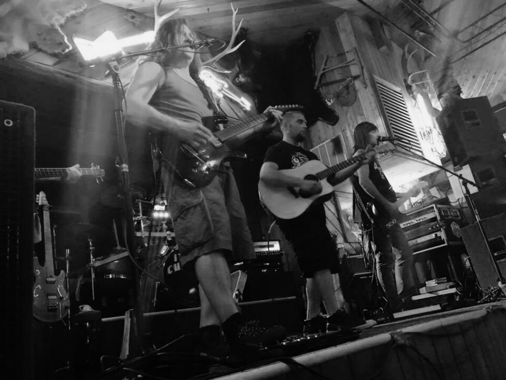 Band performing live on stage: guitarist, acoustic guitarist, and singer under stage lights.