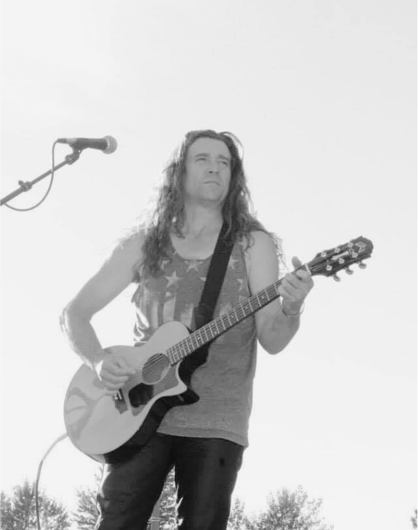Man playing an acoustic guitar, wearing a stars-and-stripes tank top, and standing near a microphone outdoors.