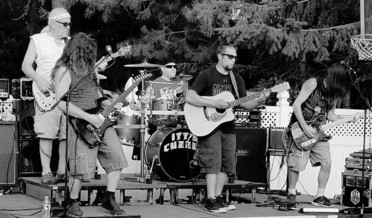 Rock band performing outdoors on a stage with instruments, members wearing sunglasses.